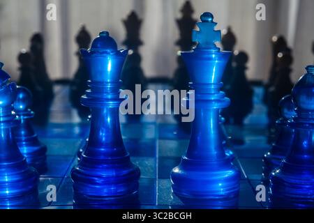 Vibrant blue chess pieces, including a king and queen, are prominently displayed on a reflective glass chessboard, with silhouettes of other pieces cr Stock Photo