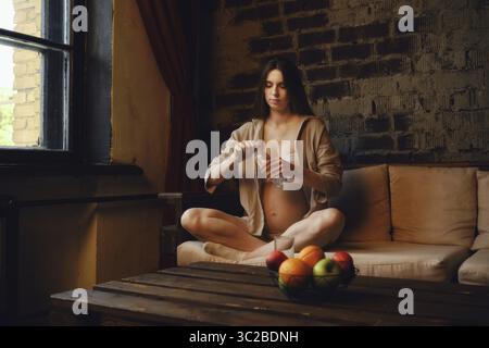 A pregnant woman sits comfortably on a sofa in her home, enjoying a moment of calm. She is focused on opening a bottle with water Stock Photo