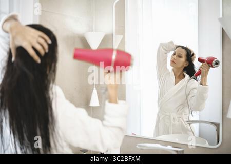 A woman is using a hair dryer to style her wet hair while standing in front of a large mirror. The bright bathroom features minimalistic design elemen Stock Photo