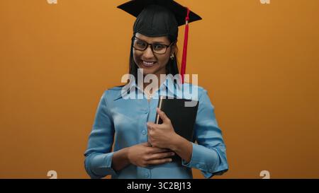 Woman graduate smiling, wearing graduation cap, holding book against vibrant orange background, embodying academic success and happiness. Stock Photo