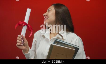 Young hispanic woman holds diploma with red ribbon and stack of books in studio; proud achievement. Stock Photo