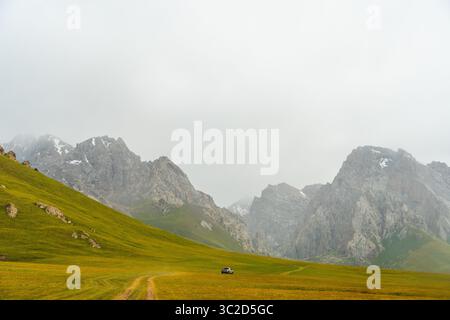 A car on a winding dirt road through expansive green and yellow mountain slopes with misty peaks in the distance Stock Photo