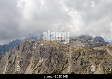 Panoramic view from Tre Cime di Lavaredo with dramatic rocky ridges, cloud-covered peaks, sparse snow, and layered Dolomite cliffs in Italy. Stock Photo