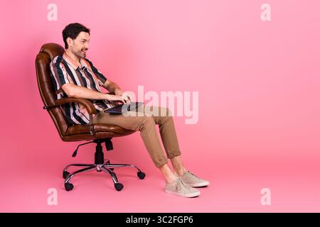 Casually dressed man engaging with keyboard while sitting in leather chair against bright pink background Stock Photo