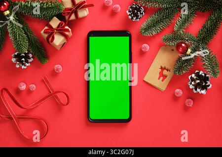 Festive Christmas smartphone mockup with green screen on red background with fir branches, pinecones, tags, ribbon and small gifts Stock Photo