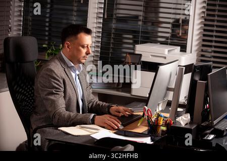 Confident businessman in a classic grey blazer focused on work at a bright office desk. Modern workspace, blinds on windows, printer and monitors. Stock Photo