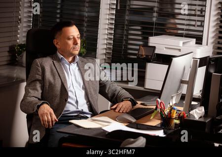 Pensive businessman in a blazer sitting at desk in modern office, staring thoughtfully at computer screen. Calm expression, workplace with supplies. Stock Photo