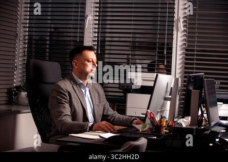 Confident businessman in a classic grey blazer focused on work at a bright office desk. Modern workspace, blinds on windows, printer and monitors. Stock Photo