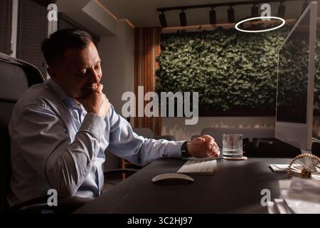 Smiling middle-aged businessman relaxing at his desk in a modern eco-friendly office. Warm light, stylish interior, and casual mood. Stock Photo