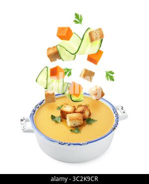 Croutons, cucumber slices and pumpkin cubes falling into bowl of soup against white background Stock Photo