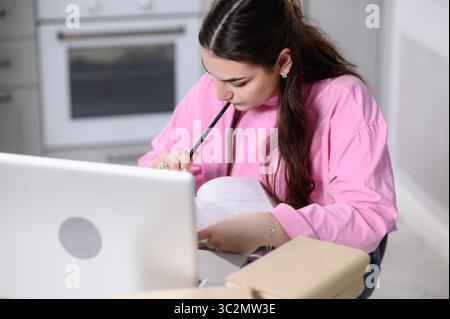 Woman sits at table with laptop, looks at notes in notebook, holding pen to lips, writing poetry, doing homework. Girl writes down desires, goals in diary, bringing pen to lips, thinking in kitchen Stock Photo