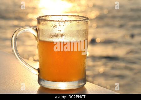 A chilled Mexican beer glass captures the warm glow of a coastal sunset with soft sea bokeh in the background. Stock Photo