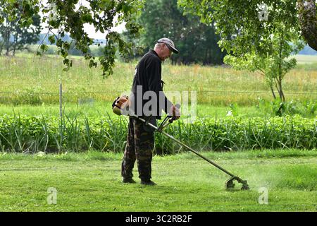 Old man using grass trimmer in backyard field during sunny day. Wearing dark clothes and cap, surrounded by trees and wild grass. Rural lifestyle and Stock Photo