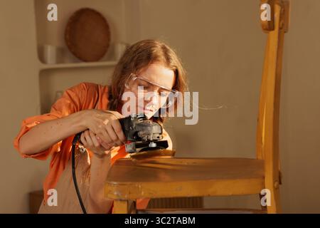 Woman using electric sander on wooden chair, home furniture restoration, repair and sanding process. Stock Photo