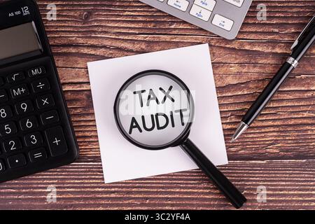 Magnifying glass focusing on the words "TAX AUDIT" with a calculator, pen, and keyboard on a wooden desk. Stock Photo