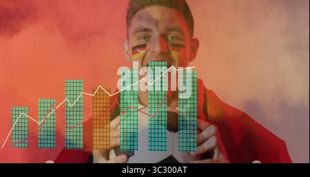 Shouting sports fan holding soccer ball in studio, with red cape, flag face paint, data charts Stock Photo