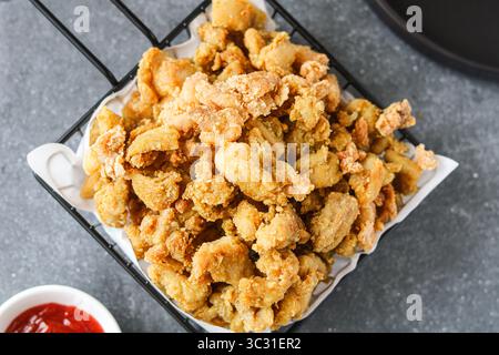 Top view of golden-brown popcorn chicken or bite-sized fried chicken pieces, piled high in a black wire serving basket lined with white paper. Stock Photo