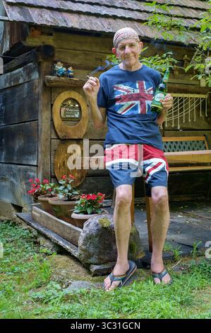 smiling older man wearing union flag shorts t.shirt in rural countryside garden barbecue holding bottle of soproni lager beer drink with cigarette Stock Photo