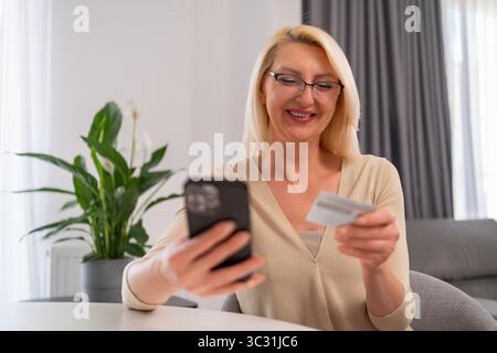 Smiling blonde woman using her smartphone to shop online and pay with a credit card at home Stock Photo