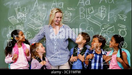 Hugging teacher holding students chatting in classroom, with chalkboard doodles and backpacks Stock Photo