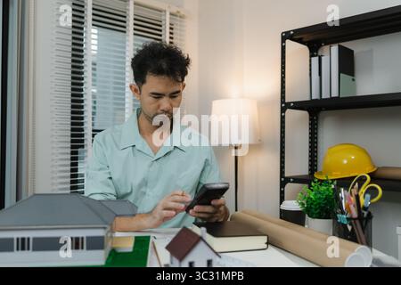 Asian male architect working with a calculator at his desk in a modern office. Surrounded by house models, tools, and books, he focuses on constructio Stock Photo