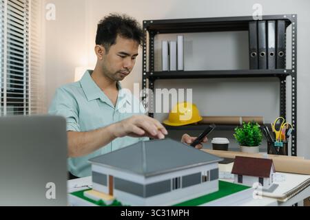 Asian male architect working with a calculator at his desk in a modern office. Surrounded by house models, tools, and books, he focuses on constructio Stock Photo