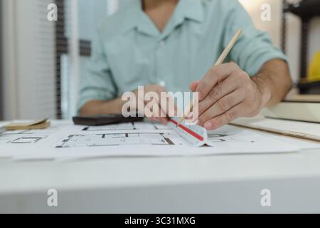 An architect working on house plans at a white desk with blueprints, a calculator, and a triangular scale ruler, focusing on drawing and measuring det Stock Photo