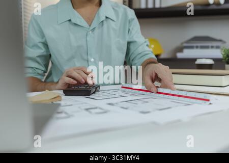 An architect working on house plans at a white desk with blueprints, a calculator, and a triangular scale ruler, focusing on drawing and measuring det Stock Photo