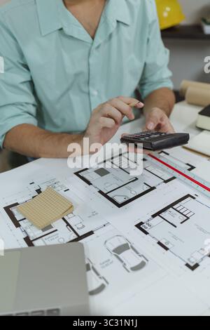An architect in a green shirt is sitting at a white desk, using a calculator while reviewing printed house floor plans. The image highlights the works Stock Photo