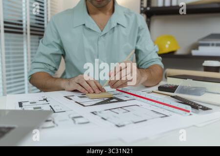 Architect working at desk with architectural blueprints, holding a pencil for sketching or adjustments. Professional workspace with tools and equipmen Stock Photo