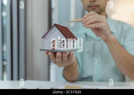 A man holding a small house model with one hand while pointing at the roof with a pencil. The scene conveys architectural design, planning, or real es Stock Photo