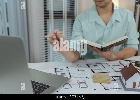 Architect working on house design project using a laptop, holding a notebook and pencil. Floor plans and a small house model are visible on the table, Stock Photo