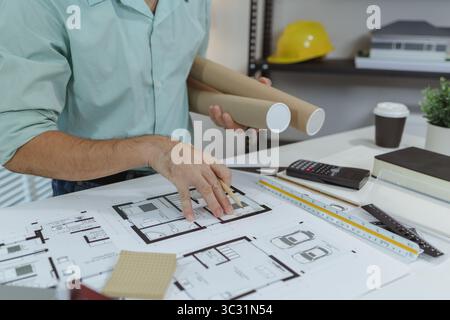 An architect pointing at house floor plans with a pencil while holding blueprint rolls. The desk is filled with architectural tools, scale ruler, and Stock Photo
