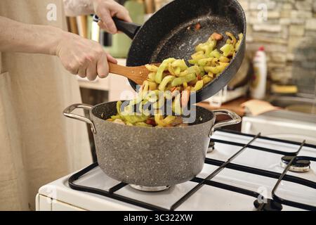 A person transfers sliced vegetables in a pot while adding more ingredients from a frying pan on a gas stove. The kitchen features warm colors and a h Stock Photo