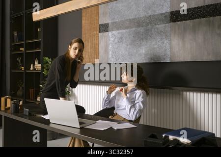 Two women engage in a conversation at a sleek office desk. One is speaking on the phone while the other listens intently Stock Photo