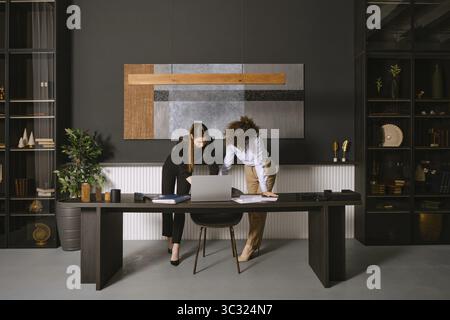 Two business professionals are engaged in a collaborative discussion around a laptop at a sleek black desk. The office features a contemporary design Stock Photo