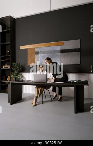 Two professionals engage in a collaborative effort at a sleek office workspace. One woman is seated at a desk, while the other stands beside her, both Stock Photo