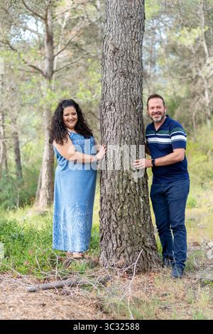 A man and woman are posing for a picture in front of a tree. The man is wearing a blue shirt and the woman is wearing a blue dress. Scene is lighthear Stock Photo