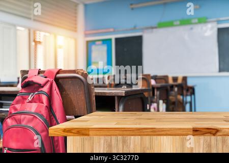 Empty classroom with wooden student desks and a red backpack hanging on a chair. Wooden table in foreground and whiteboard and bulletin board in the b Stock Photo