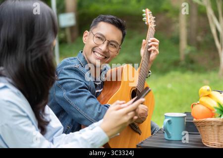 Musical Joy. Young man playing guitar while friend enjoys smartphone outdoors. Stock Photo