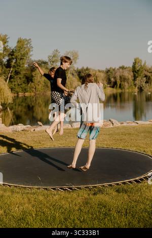 Three kids jumping together on trampoline by a calm lake Stock Photo