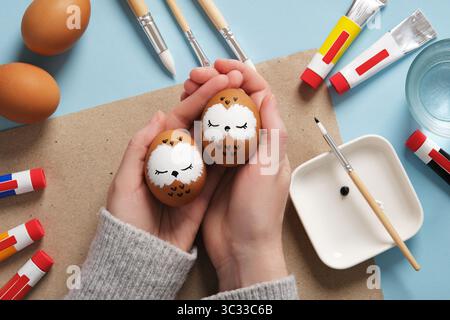 Hands holding Easter eggs with cute sleeping owls, hand-painted with acrylic paints, on a background of paints, brushes and palette. DIY concept Stock Photo