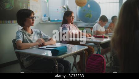 Elementary School Students Sitting at the Desks, Listening to Lecture from Teacher, Raising Hands to Provide Correct Answer. Team of Smart Diverse Kids Studying Science, Geography in Modern Classroom. Stock Photo