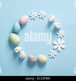 Minimalist Easter frame with white bunnies, colored eggs and wooden flowers on a blue background. Space for your text. Stock Photo