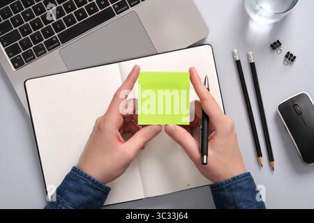 Hands holding a blank sticky note in green over an open notepad on a table. A place for your text Stock Photo