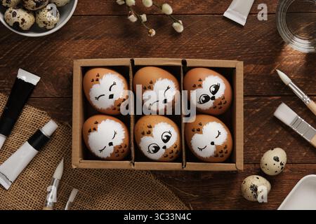 Hand-painted owl Easter eggs in a box. Surrounded by painting supplies on a wooden background. Creative holiday decoration Stock Photo