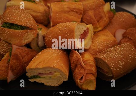 Assorted sandwiches in a black platter, viewed from above. Filled with cheese, lettuce, and deli meats in sesame rolls and croissants. Stock Photo