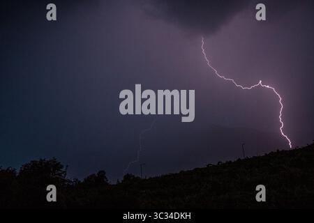 Lightning strike captured over a forested mountain ridge in Trentino, Italy, during a summer thunderstorm. The bolt illuminates the night sky in thisl Stock Photo