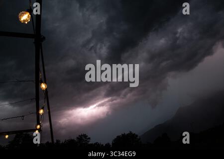 Dark storm clouds rolling over mountain peaks in Trentino, Italy, during a severe summer thunderstorm. Warm lights from outdoor string bulbs contrast Stock Photo