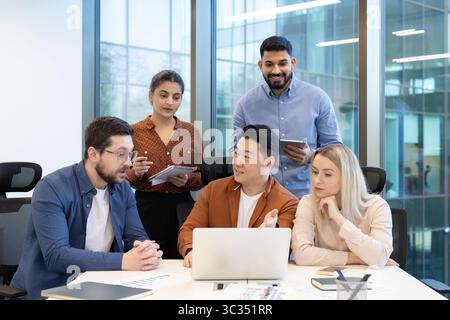 A diverse team collaborates around a laptop in a modern office, discussing ideas and strategies. Stock Photo